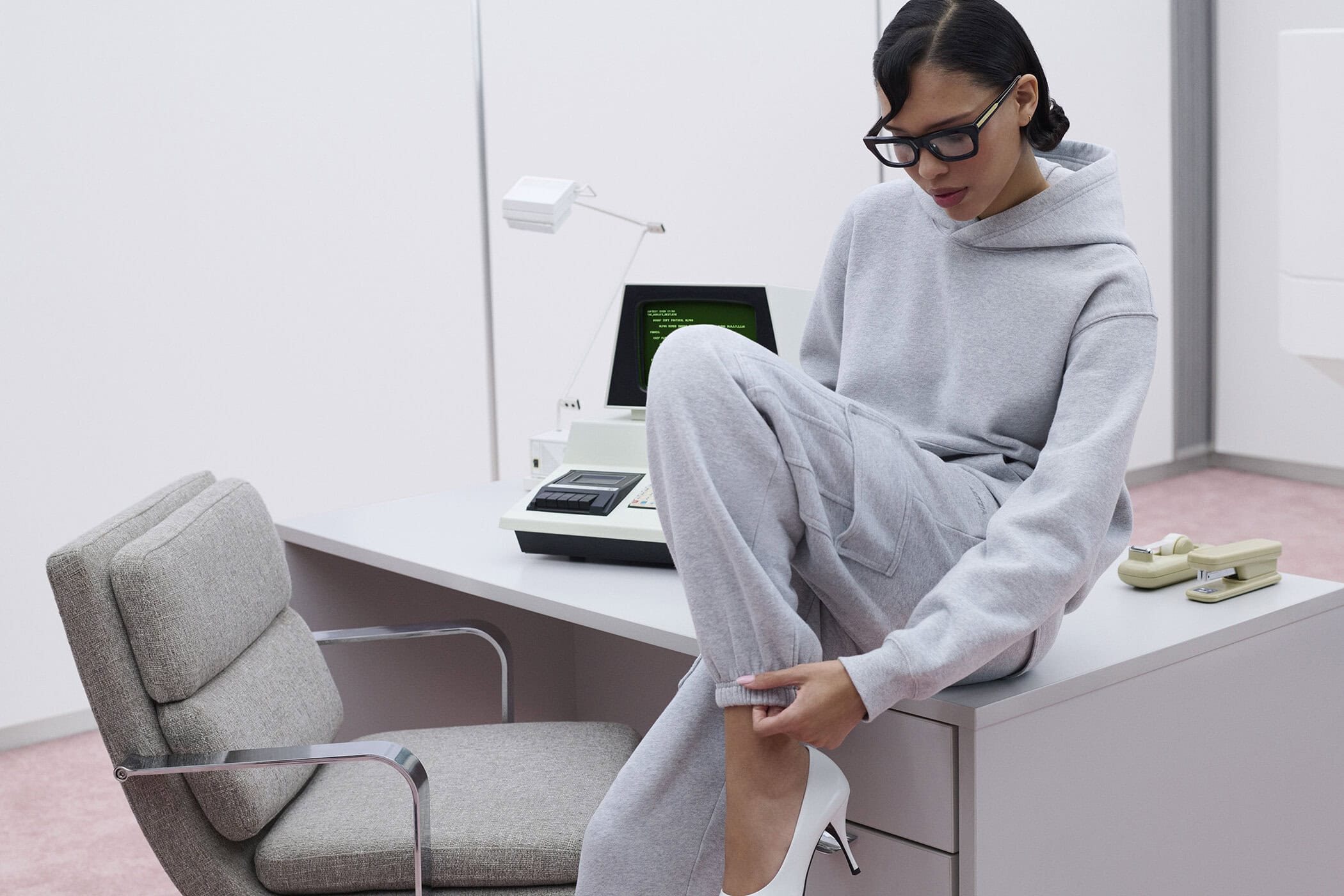 Model Nara Aziza Smith wearing a grey sweatfleece hoodie and matching sweatpants, seated on a desk in an office.