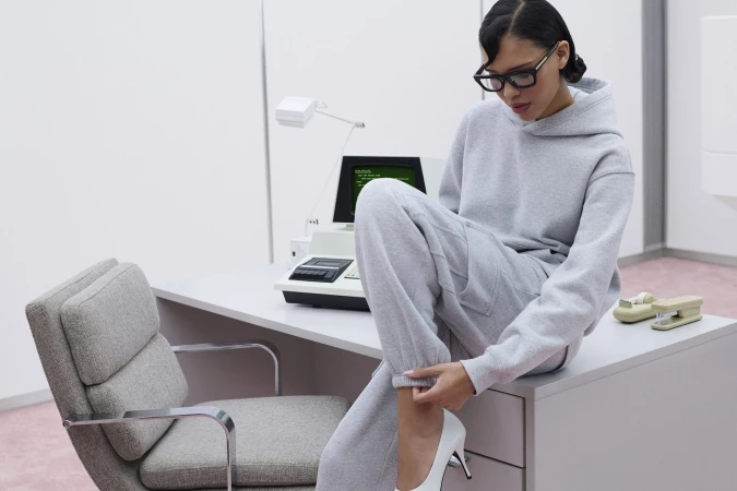Model Nara Aziza Smith wearing a grey sweatfleece hoodie and matching sweatpants, seated on a desk in an office.