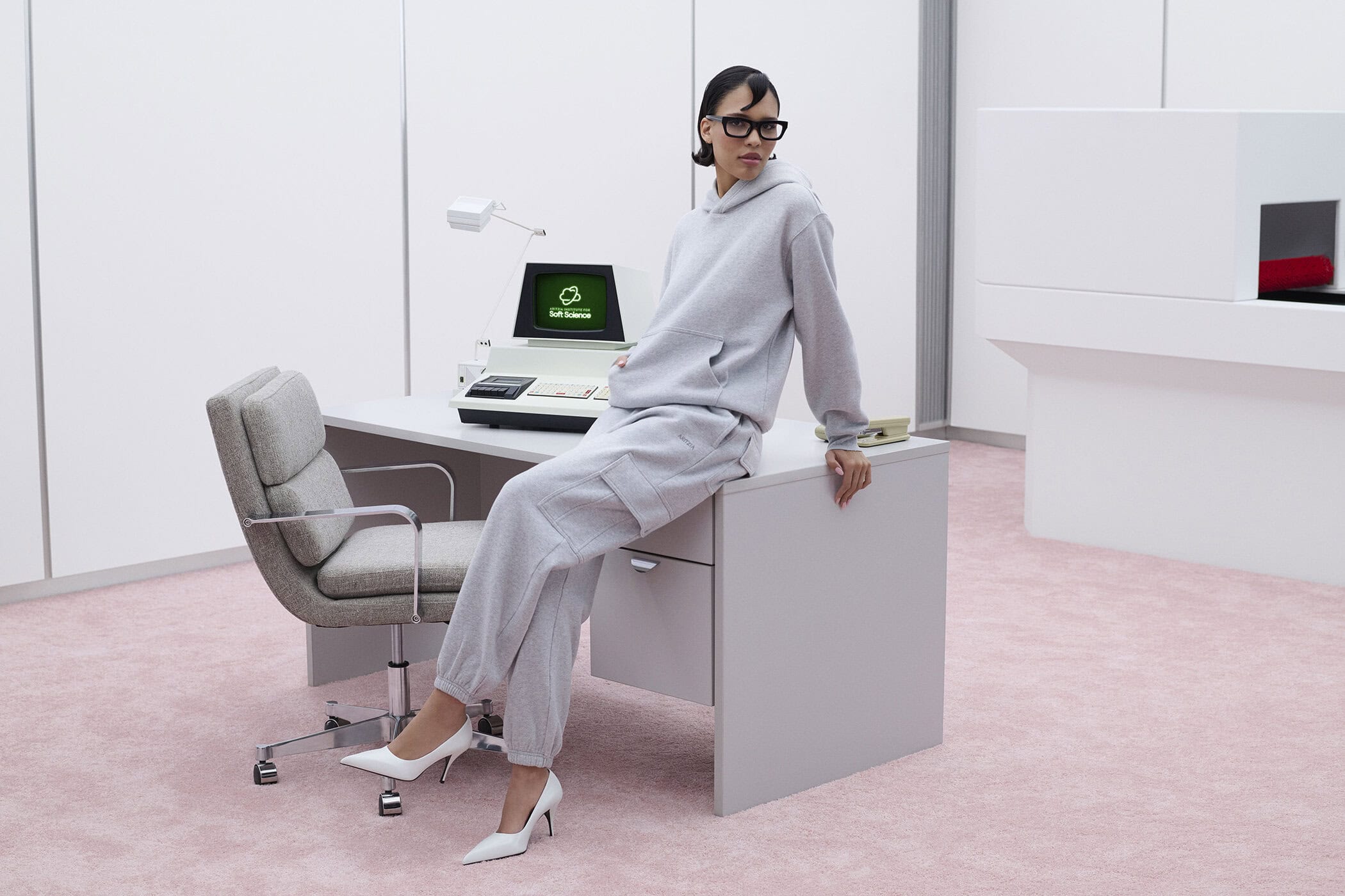 Model Nara Aziza Smith in a grey sweatfleece set sitting on an office desk in a retro-futuristic pink-carpeted room for Aritzia.