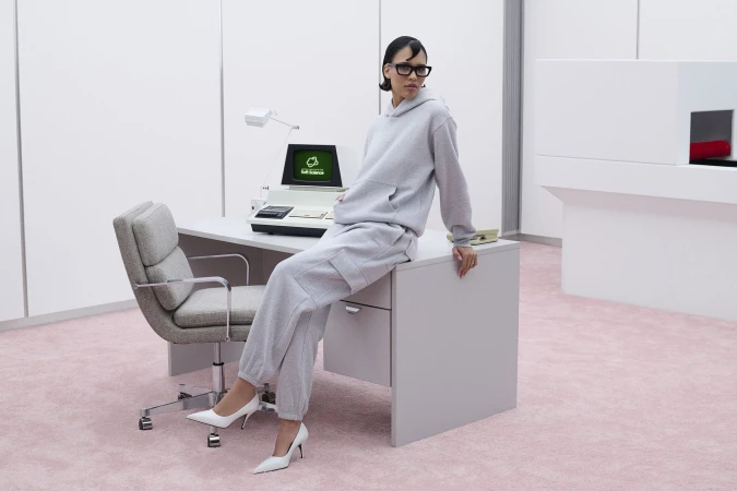 Model Nara Aziza Smith in a grey sweatfleece set sitting on an office desk in a retro-futuristic pink-carpeted room for Aritzia.