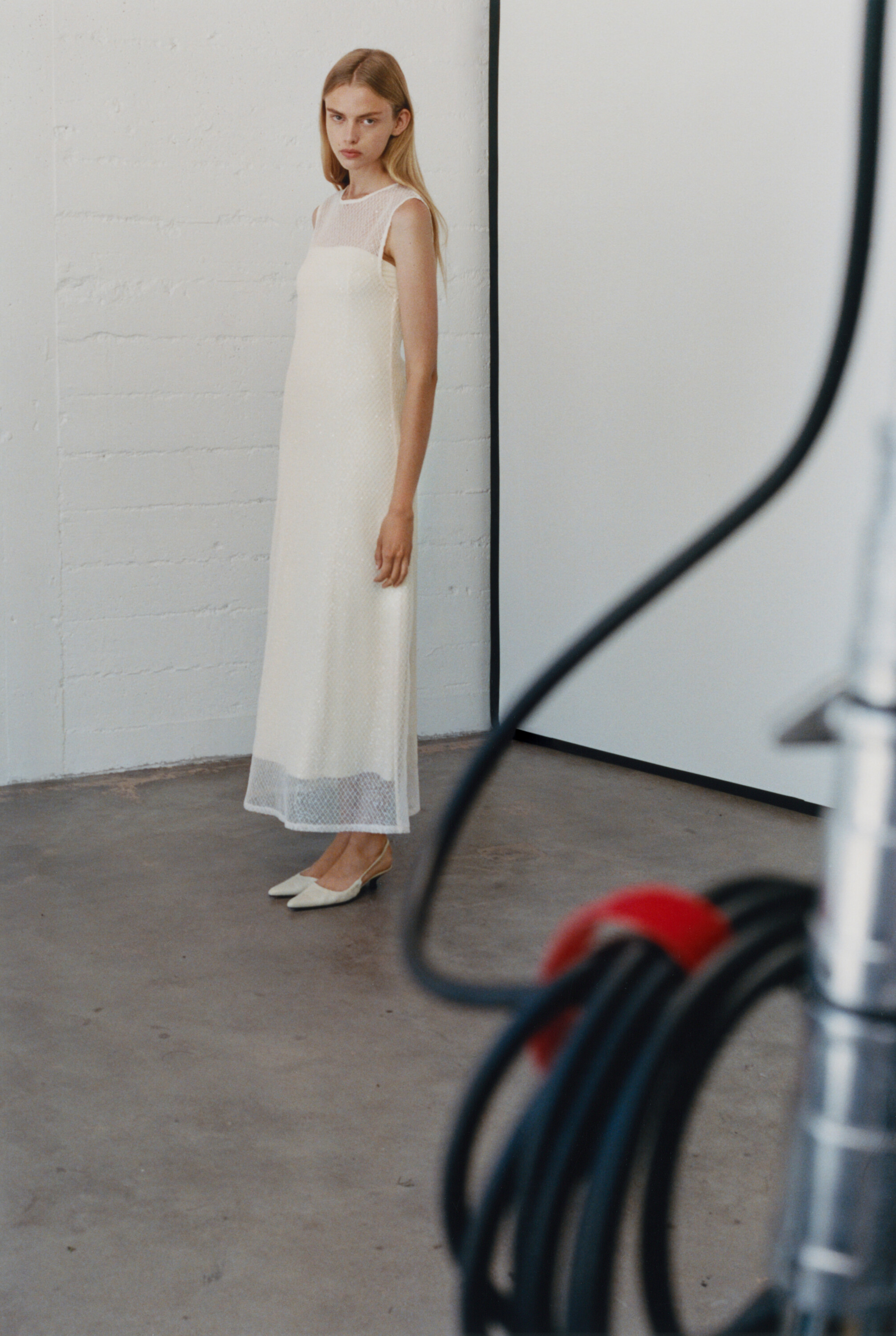 A female model wearing a long white dress stands in a studio with equipment cables in the foreground for Rue Sophie Fall 2024 campaign.