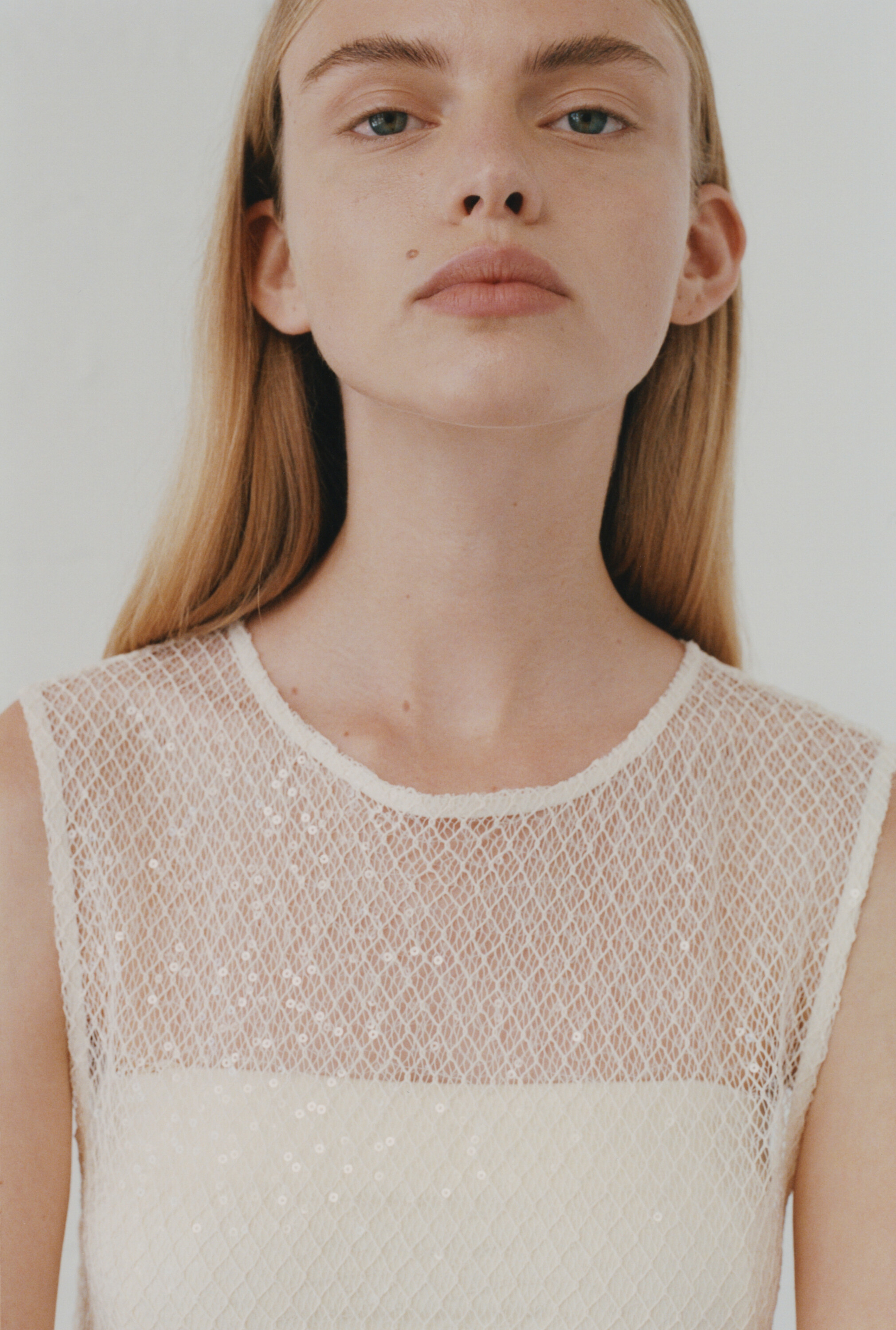 Close-up portrait of a woman wearing a sleeveless white mesh top against a plain light background