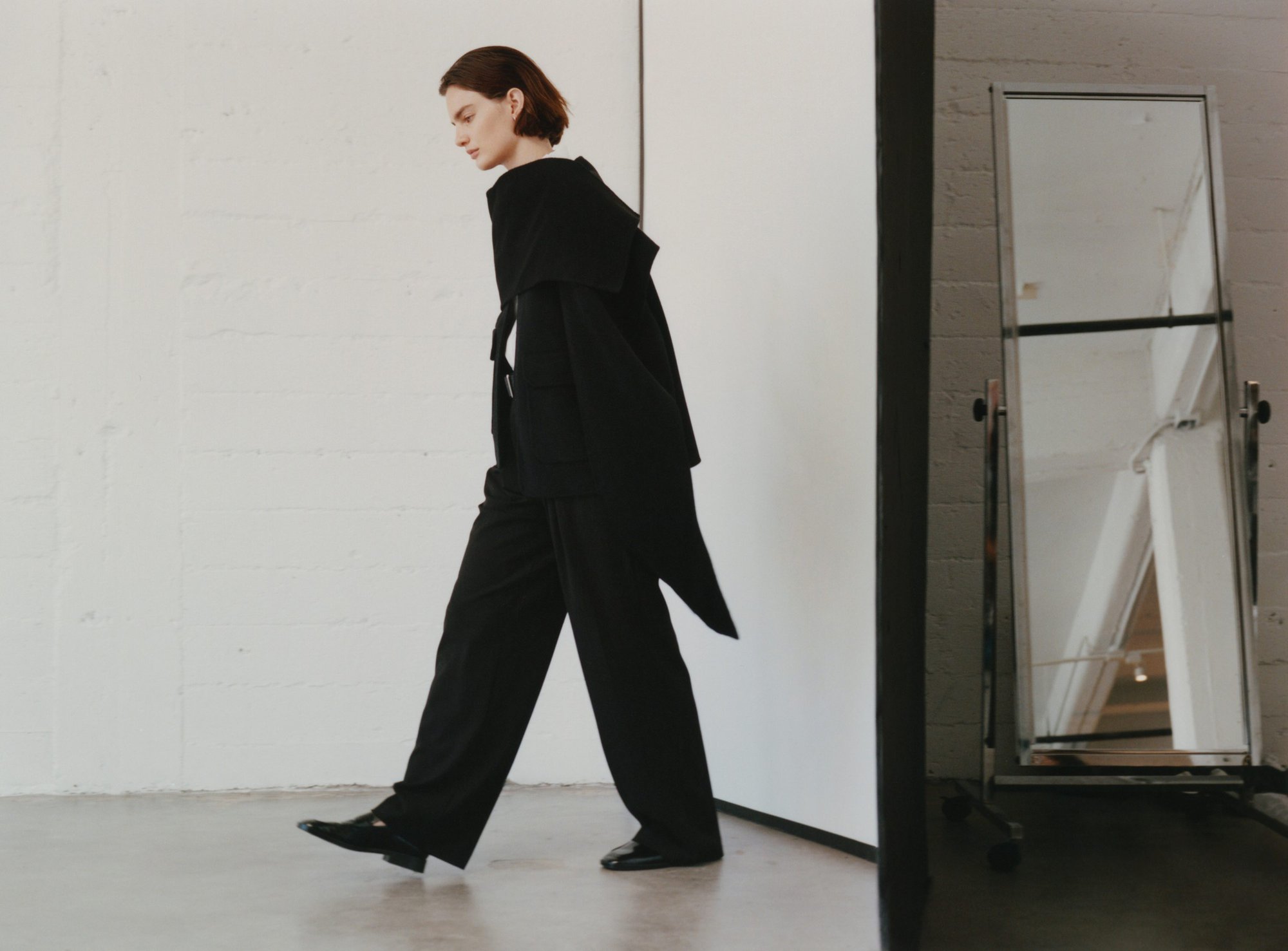 Model wearing a black coat and matching trousers walking across a light-coloured studio floor with a tall mirror in the background.