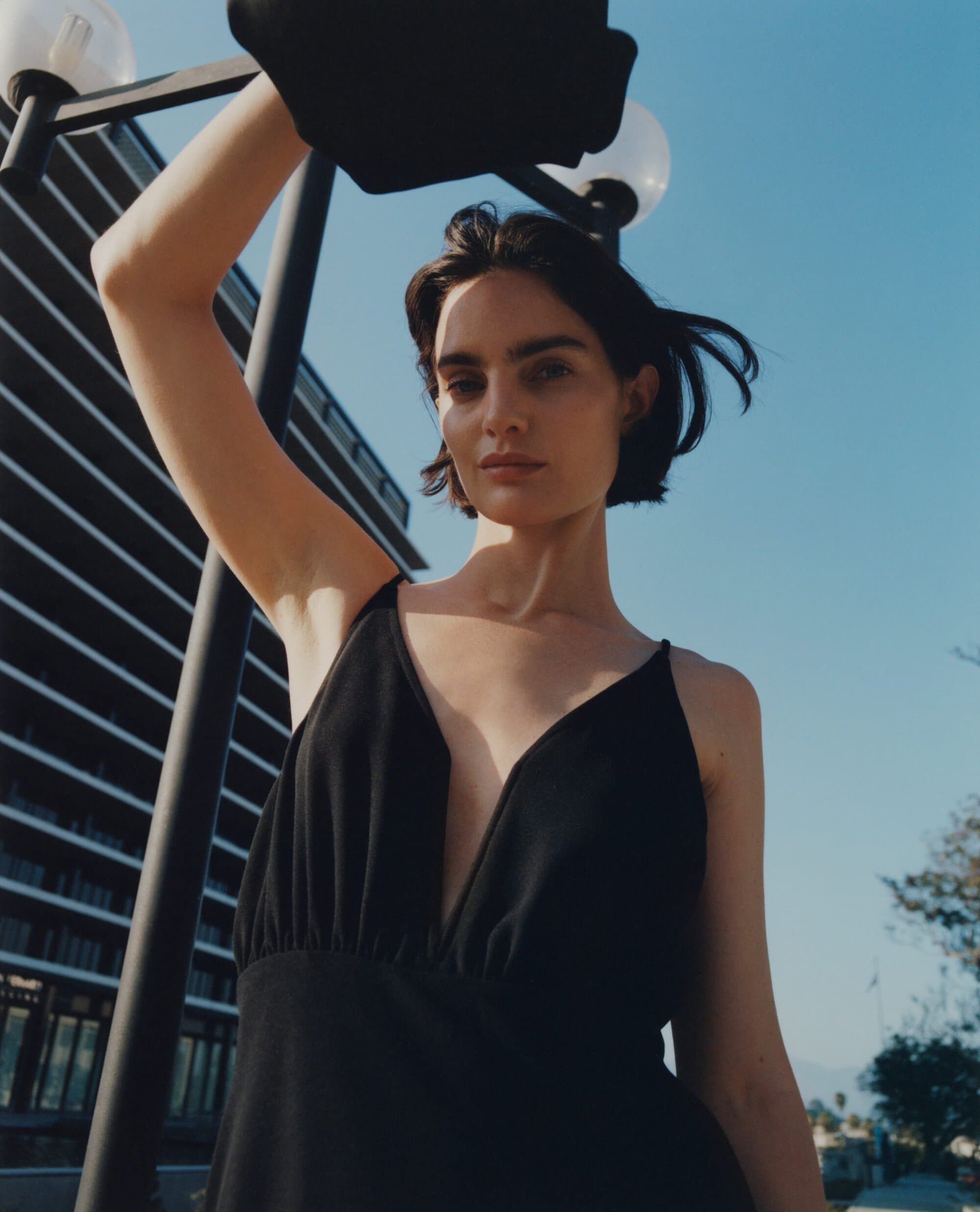 Model in a black dress raising her arms outdoors against a blue sky and modern building exterior.
