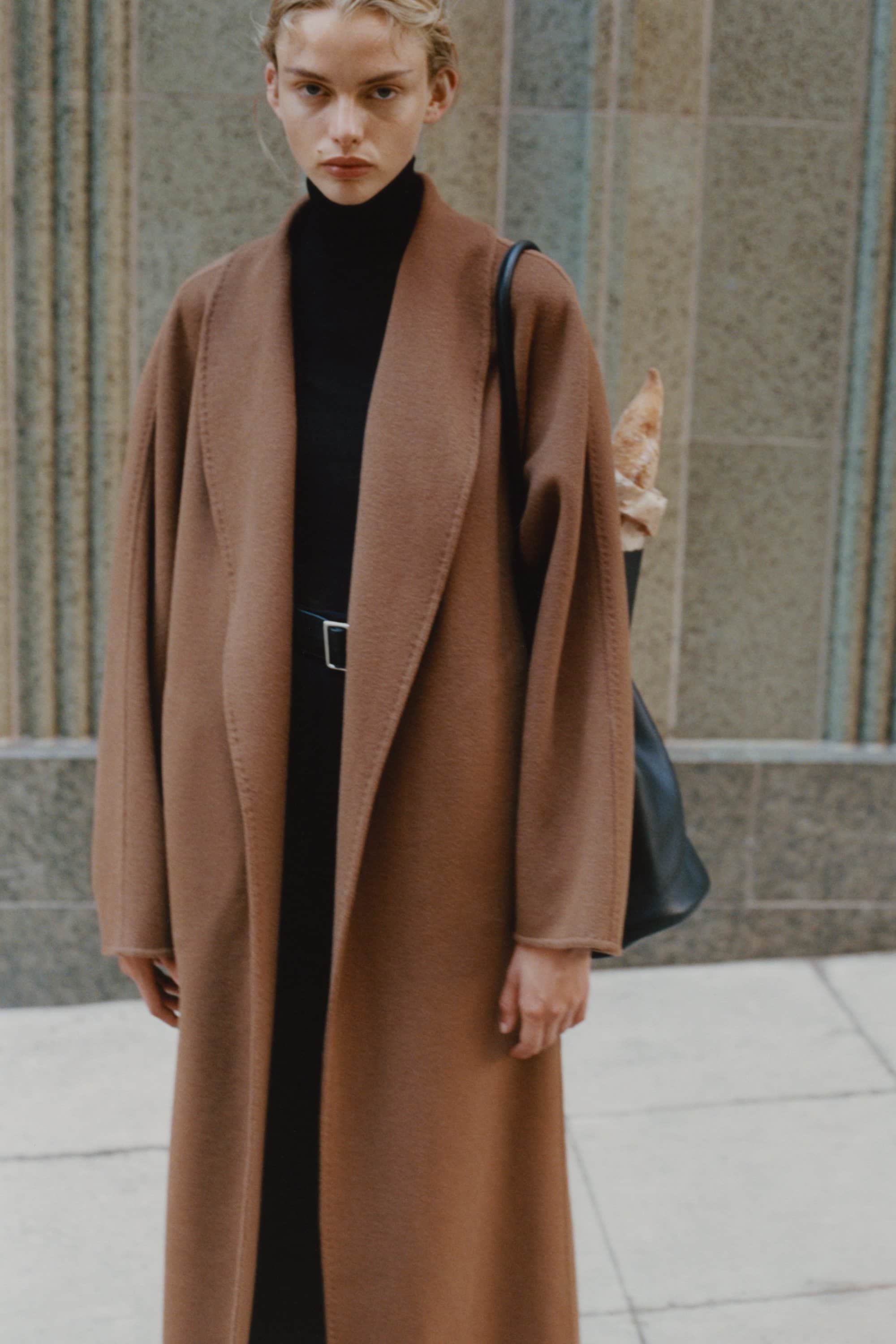 Model in a long brown coat and black turtleneck standing outdoors against stone architecture for Rue Sophie.