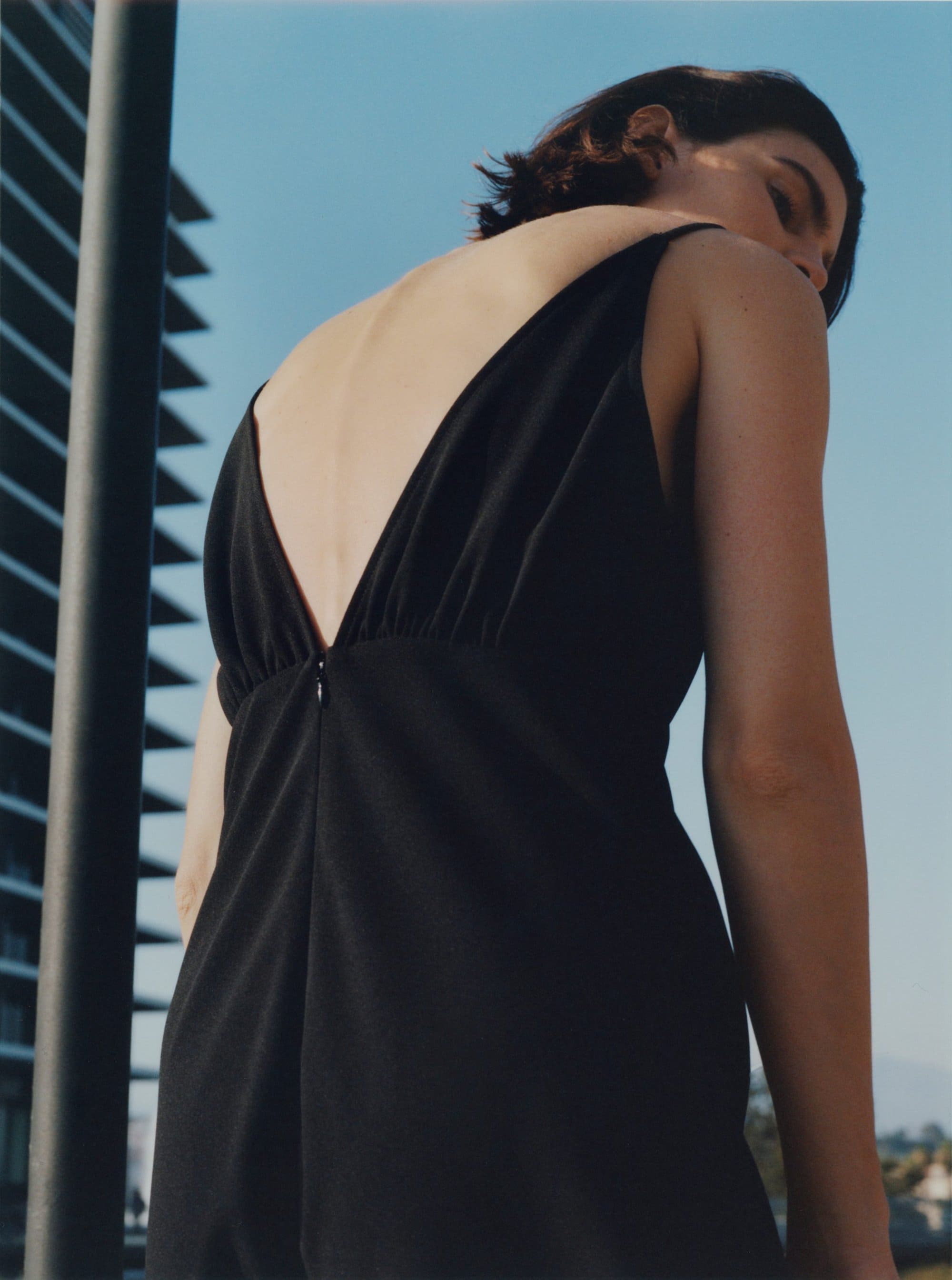 A woman in a black v-neck dress looking back against an outdoor architectural background under clear blue sky.