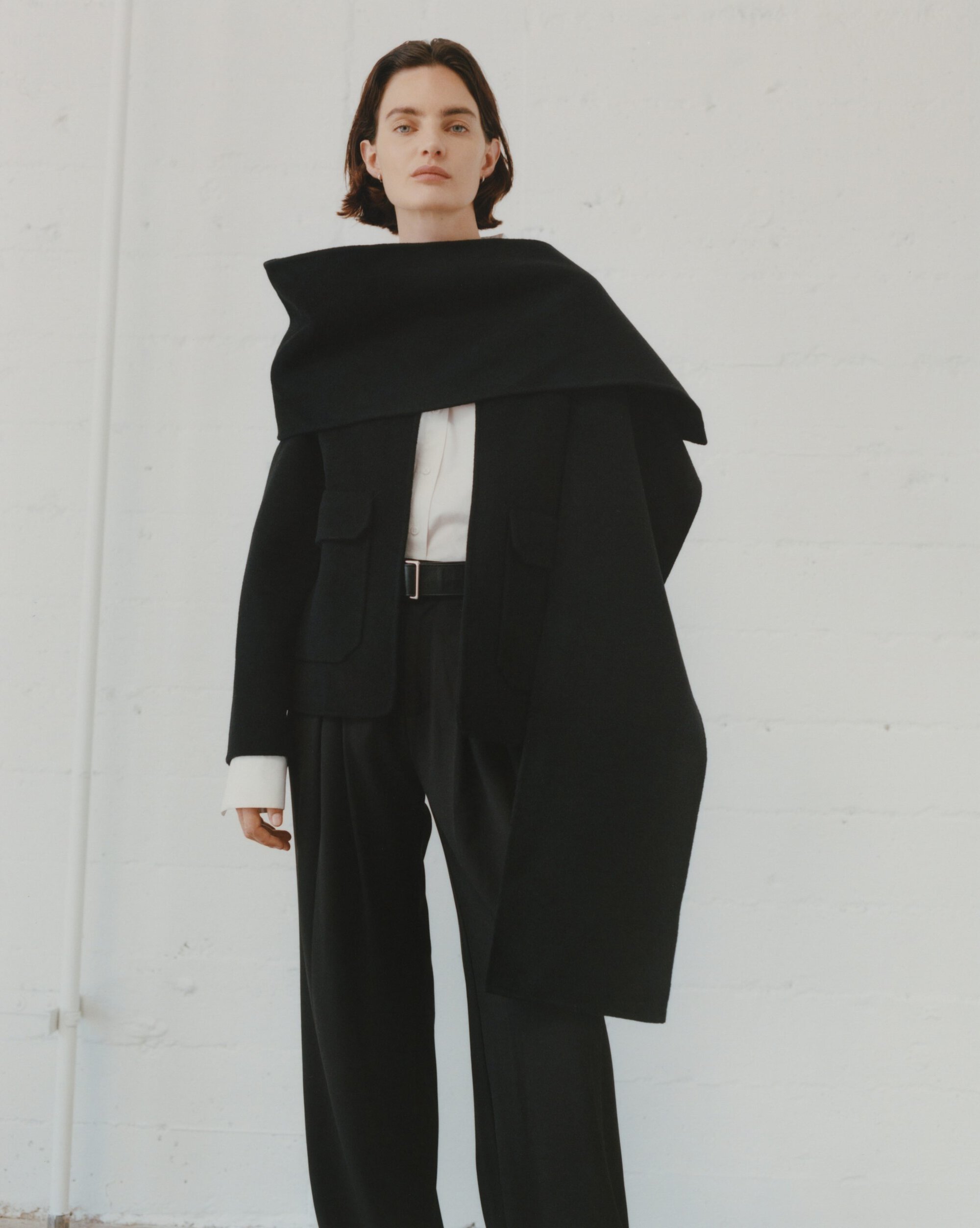 Model in black jacket with an oversized draped scarf and dark trousers standing against a white wall.