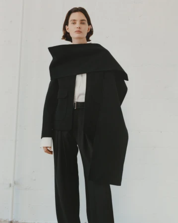 Model in black jacket with an oversized draped scarf and dark trousers standing against a white wall.