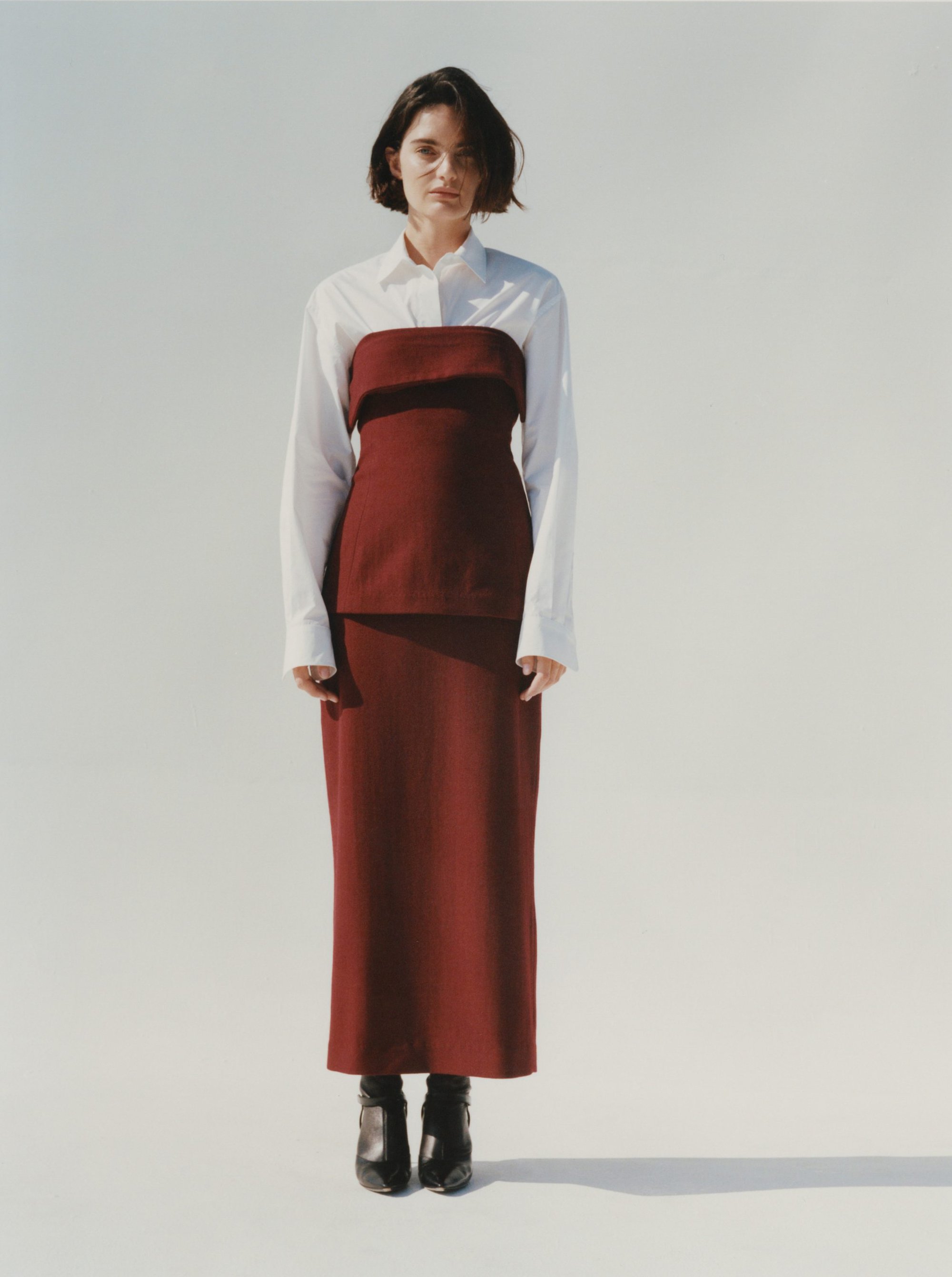 Woman with short dark hair wearing a white shirt and burgundy tube top with matching long skirt against a plain white background