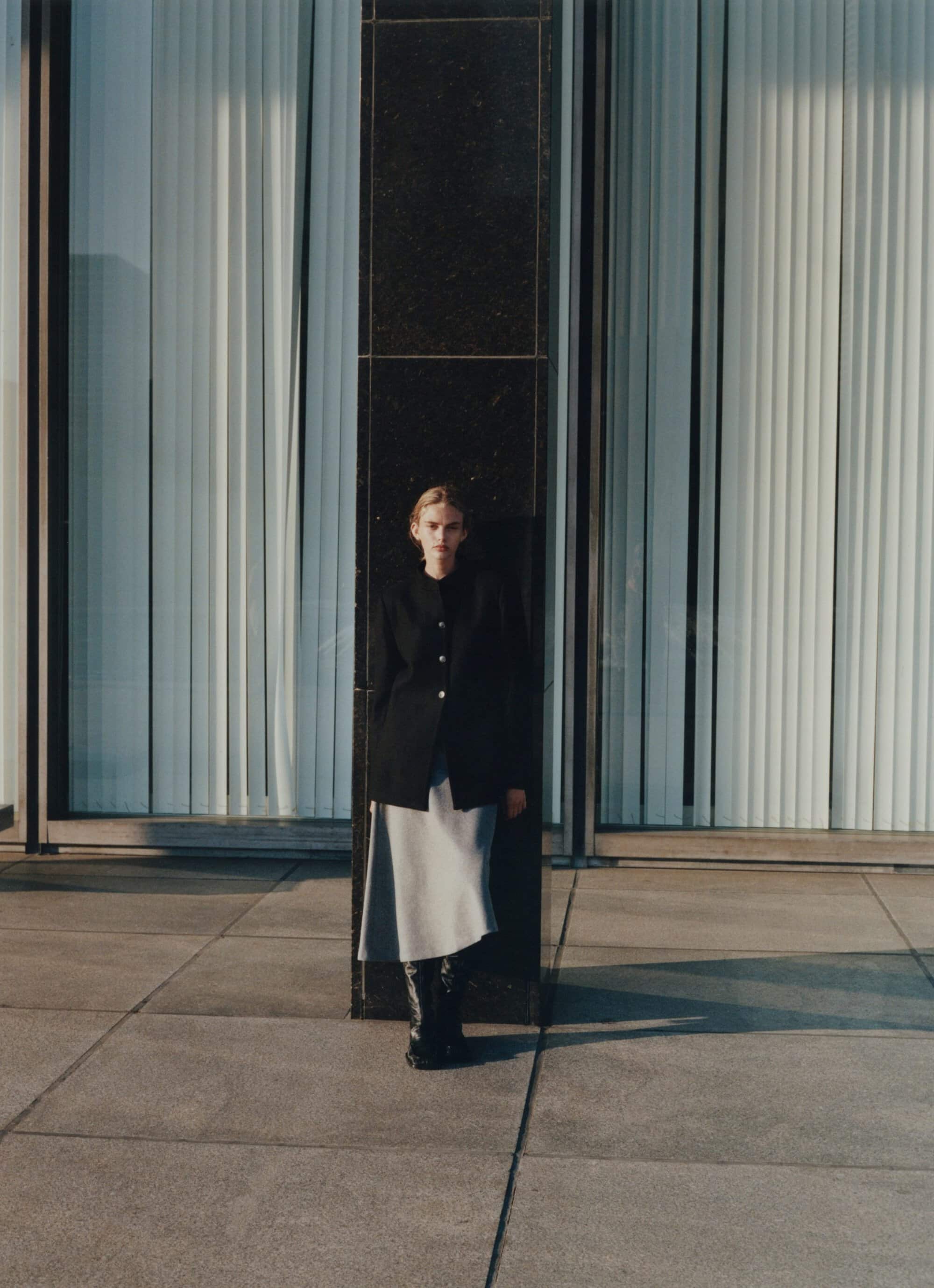 Model standing outdoors against a dark architectural pillar wearing a black jacket, grey skirt, and black boots.