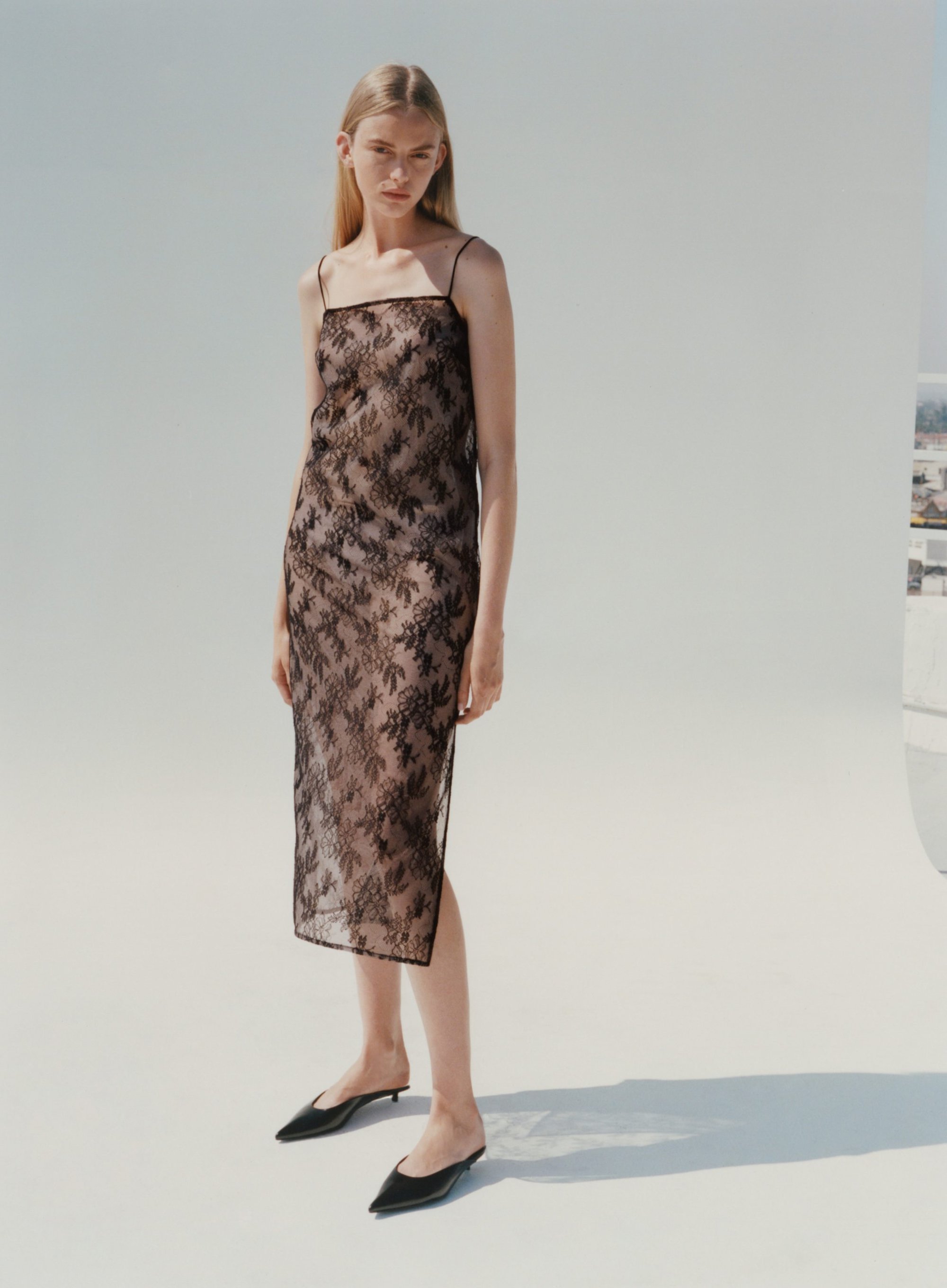 Model standing against a white seamless backdrop wearing a sheer dark lace slip dress and black mules.