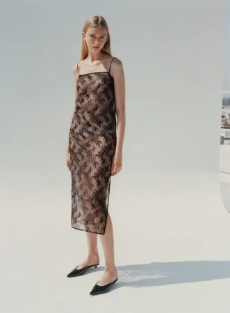 Model standing against a white seamless backdrop wearing a sheer dark lace slip dress and black mules.
