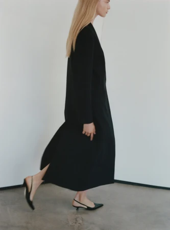 Model in profile wearing a long black coat and black slingback heels against a light wall