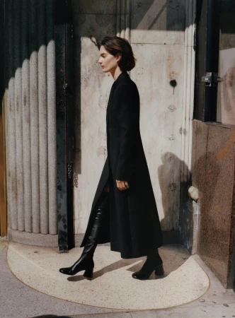 Model in a long black coat and knee-high leather boots walking past a fluted stone building exterior