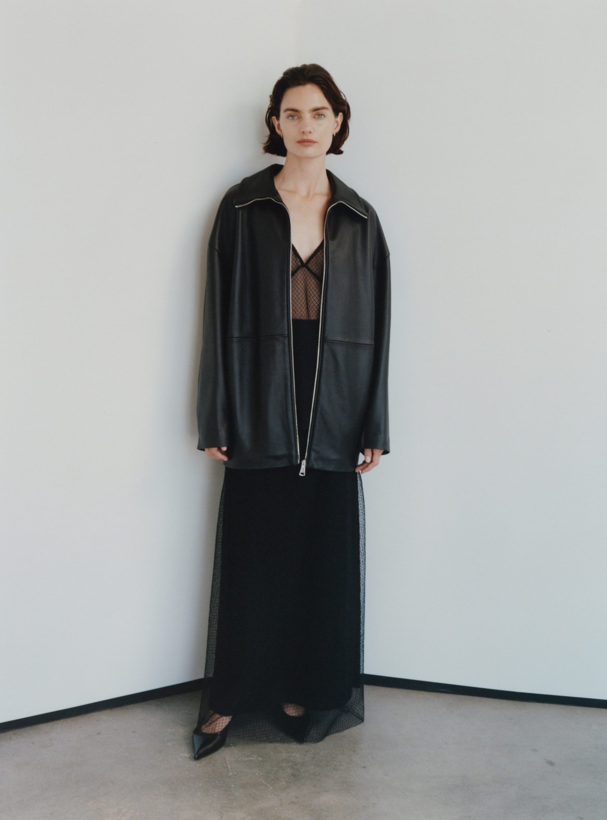 Model in black leather jacket and sheer black mesh skirt standing against a plain white wall.