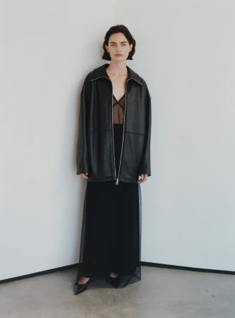 Model in black leather jacket and sheer black mesh skirt standing against a plain white wall.