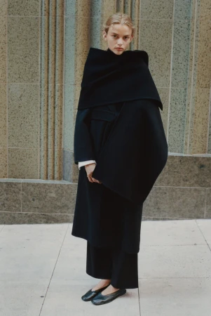 Model wearing a structured black coat and scarf standing in front of a stone building facade