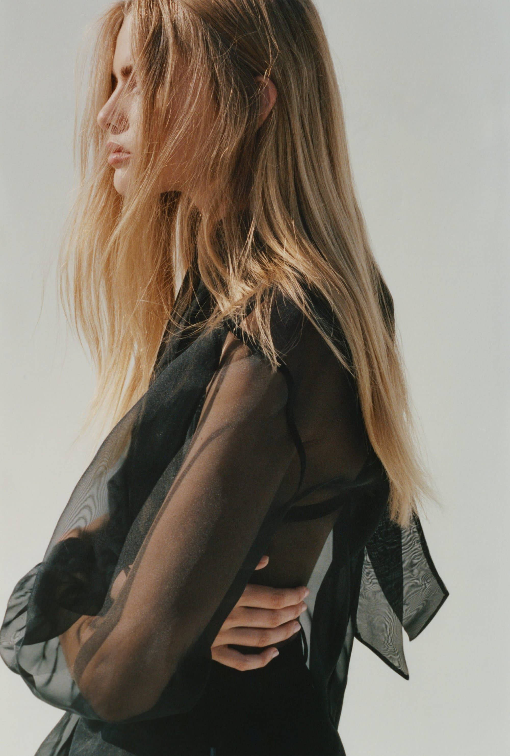 Profile view of blonde woman wearing a sheer black blouse against a plain light background