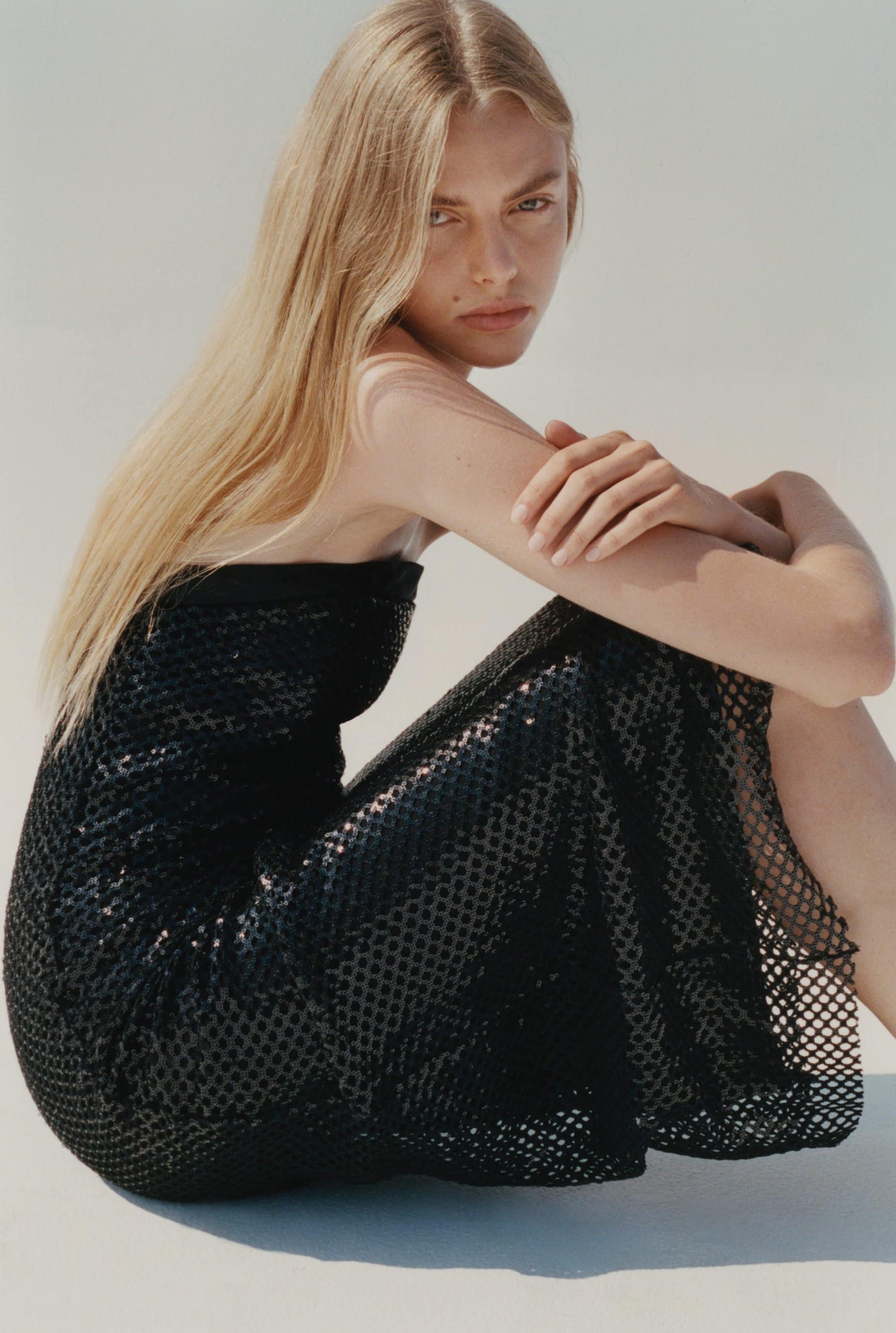 Model with long blonde hair wearing a black sequin mesh dress against a neutral white background