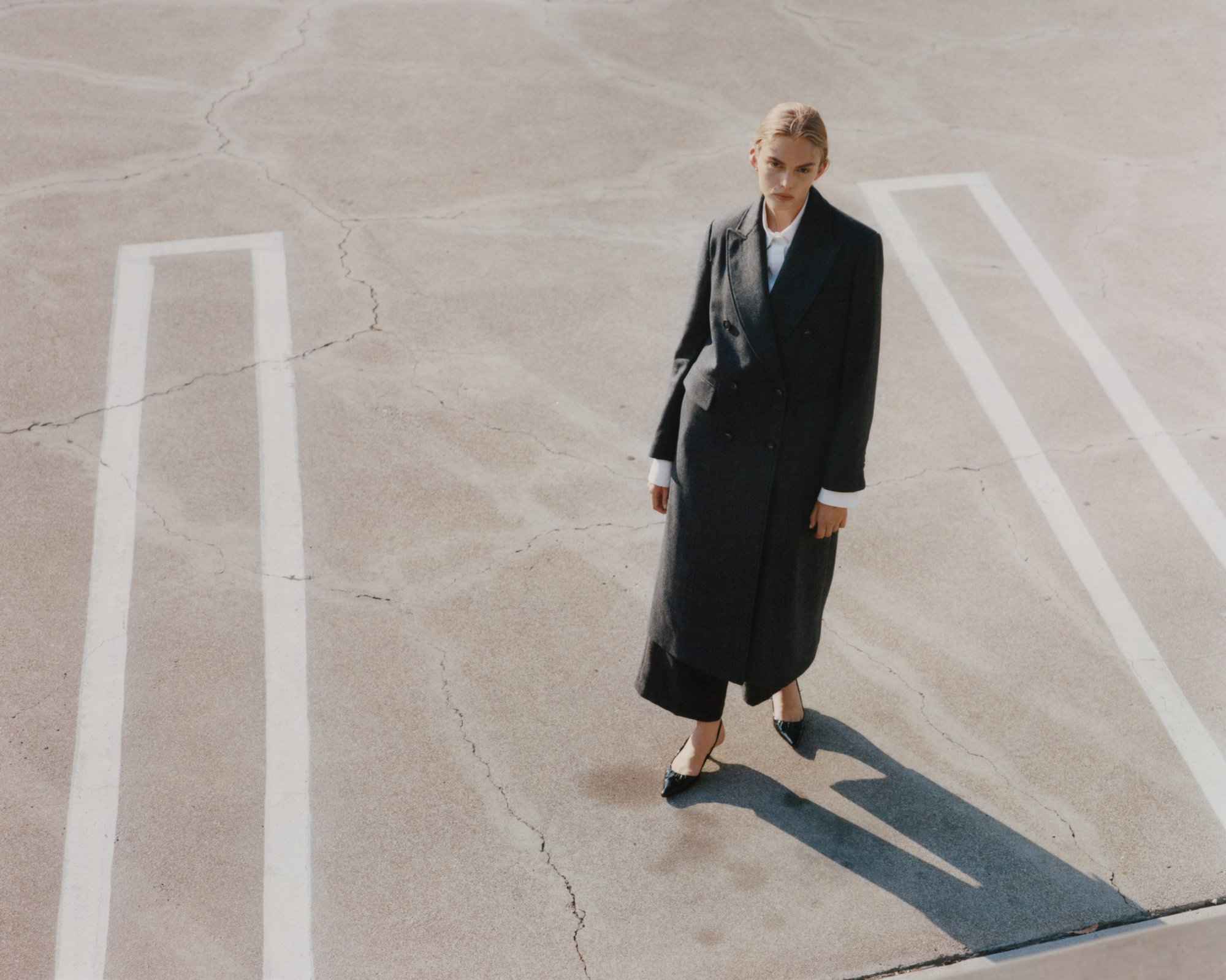 Model wearing a dark coat and heels standing on concrete pavement with white parking lines
