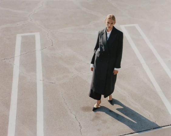 Model wearing a dark coat and heels standing on concrete pavement with white parking lines