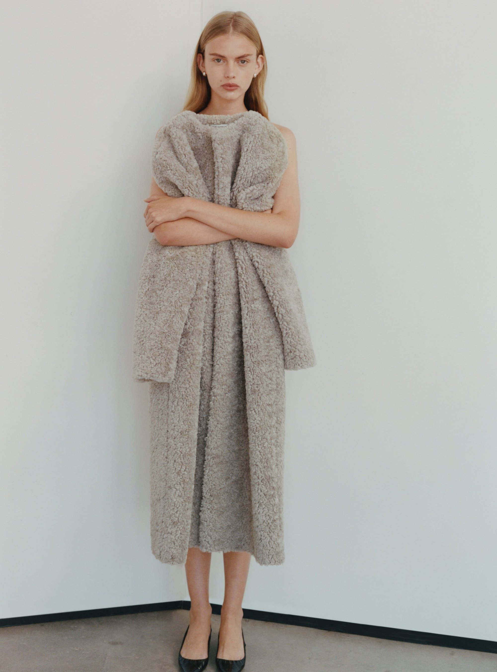 Model standing against a plain white background wearing a textured grey coat with crossed arms