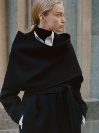 Model with blonde hair tied back wearing a black coat and white shirt against an architectural background