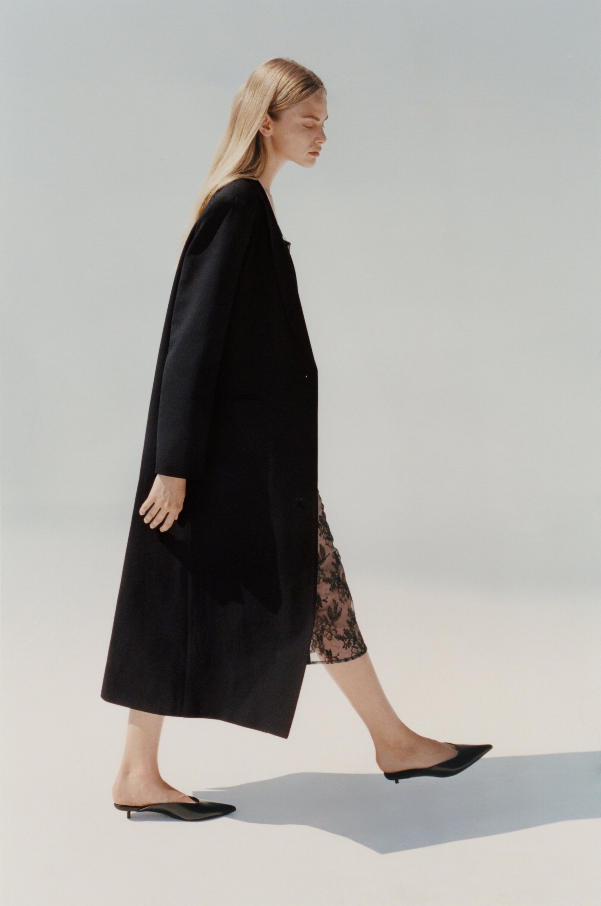 Model in a long black coat and pointed-toe flats walking across a plain light studio background.