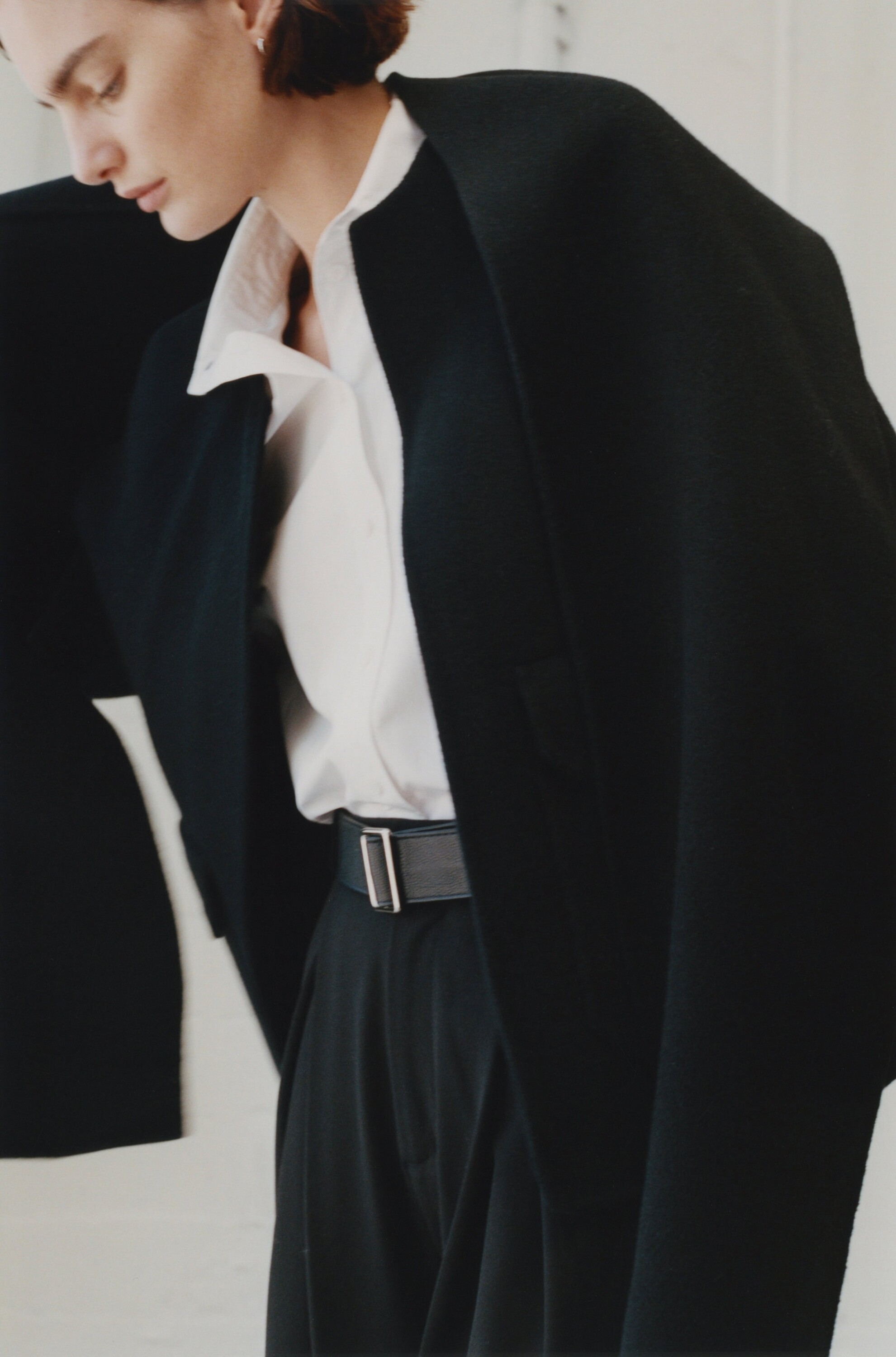 Woman in a white collared shirt and black trousers wearing a structured black coat draped over her shoulders