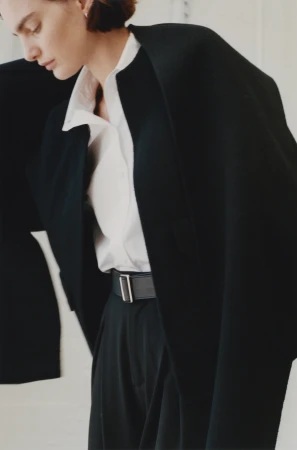 Woman in a white collared shirt and black trousers wearing a structured black coat draped over her shoulders