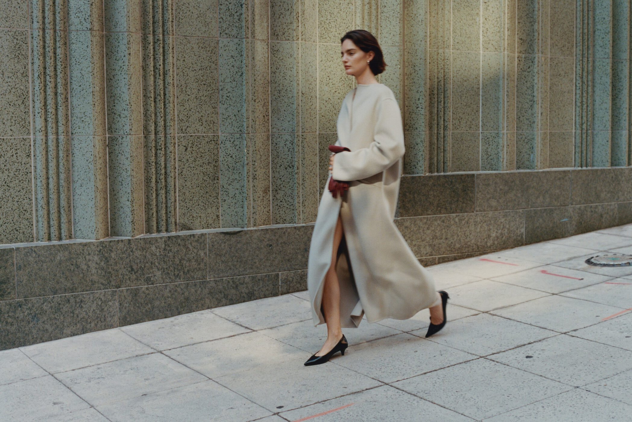 Model in a long beige coat and black heels walking past a stone building exterior