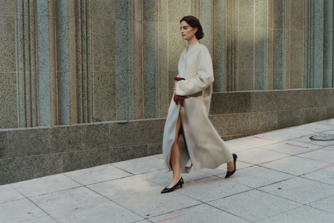 Model in a long beige coat and black heels walking past a stone building exterior