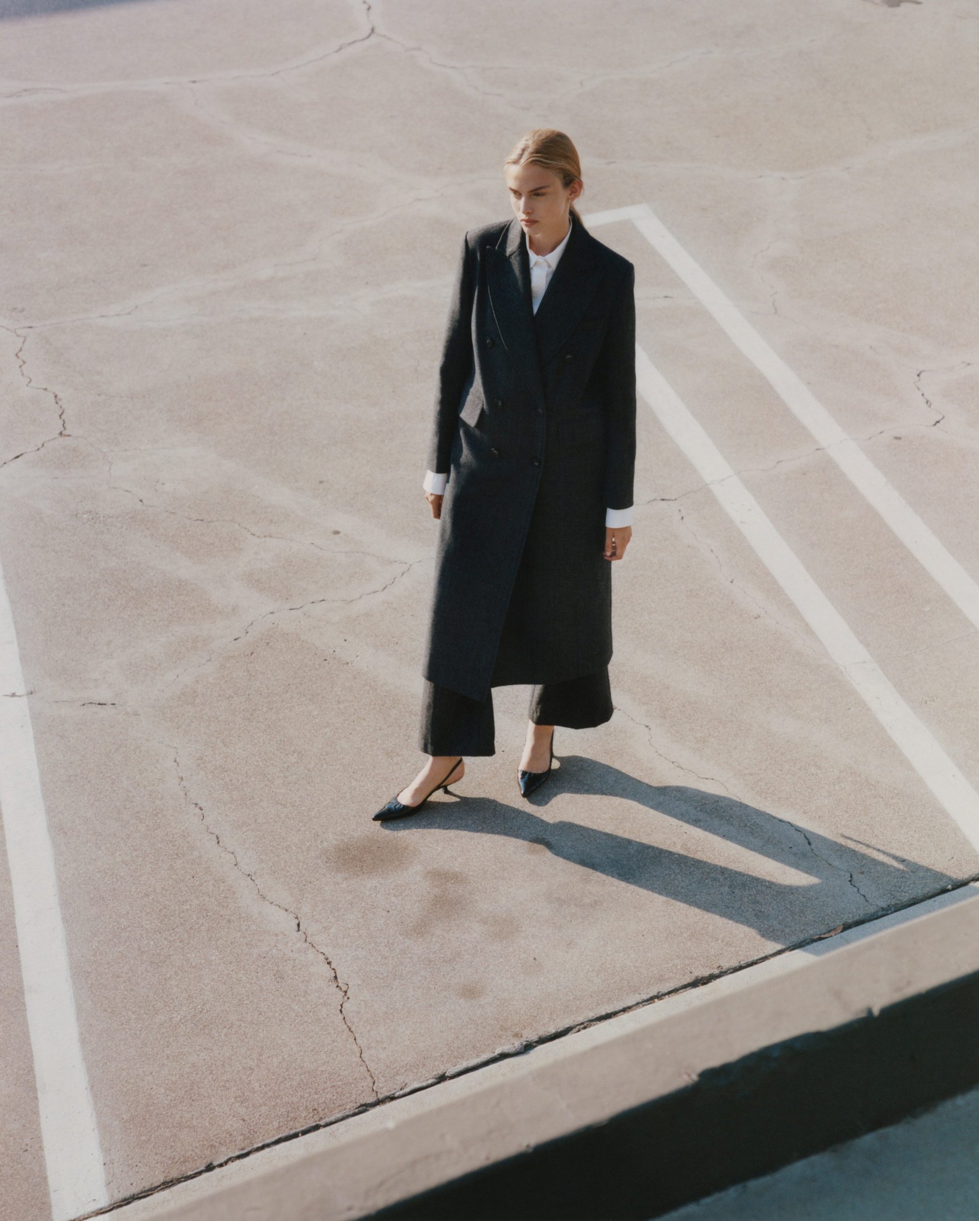 Model standing outdoors wearing a long black coat and heels on a paved surface with white painted lines
