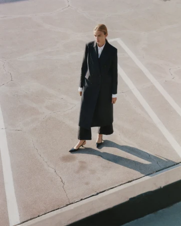 Model standing outdoors wearing a long black coat and heels on a paved surface with white painted lines