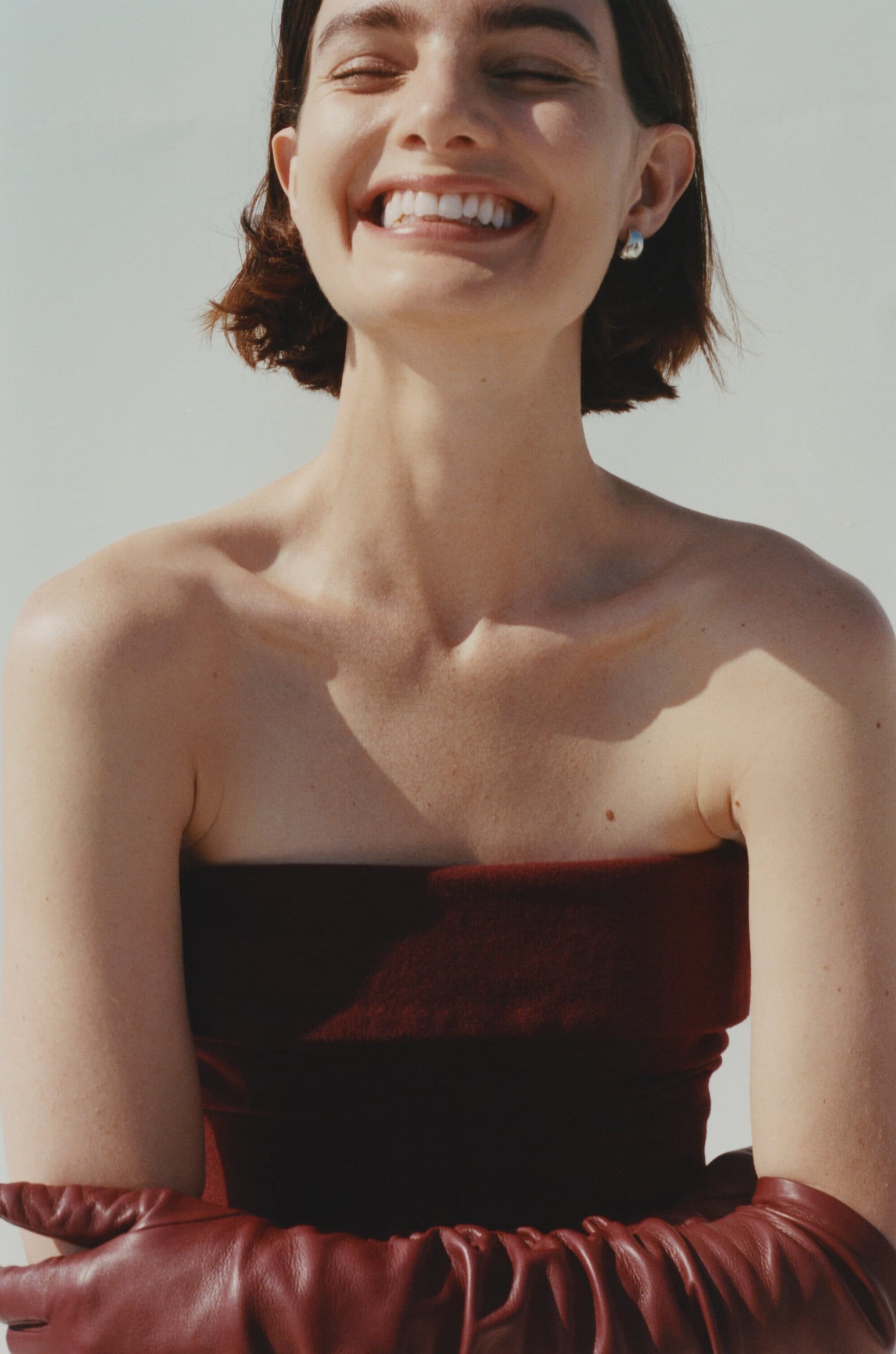 Close-up of a smiling woman wearing a strapless burgundy top and matching long leather gloves against a white wall.