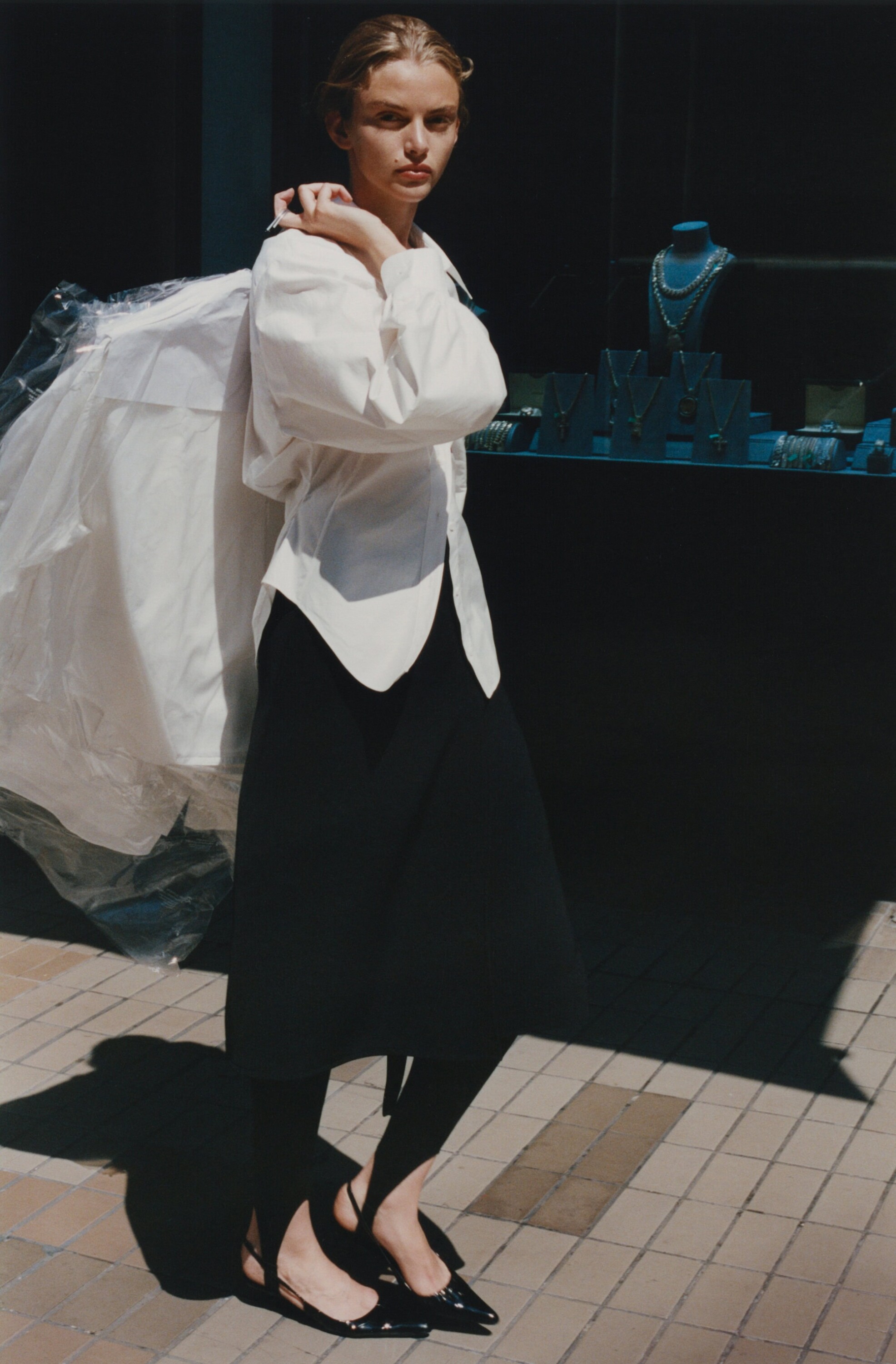 Model in white shirt and black skirt carrying a garment bag outdoors by a shop window for Rue Sophie Fall 2024 campaign.