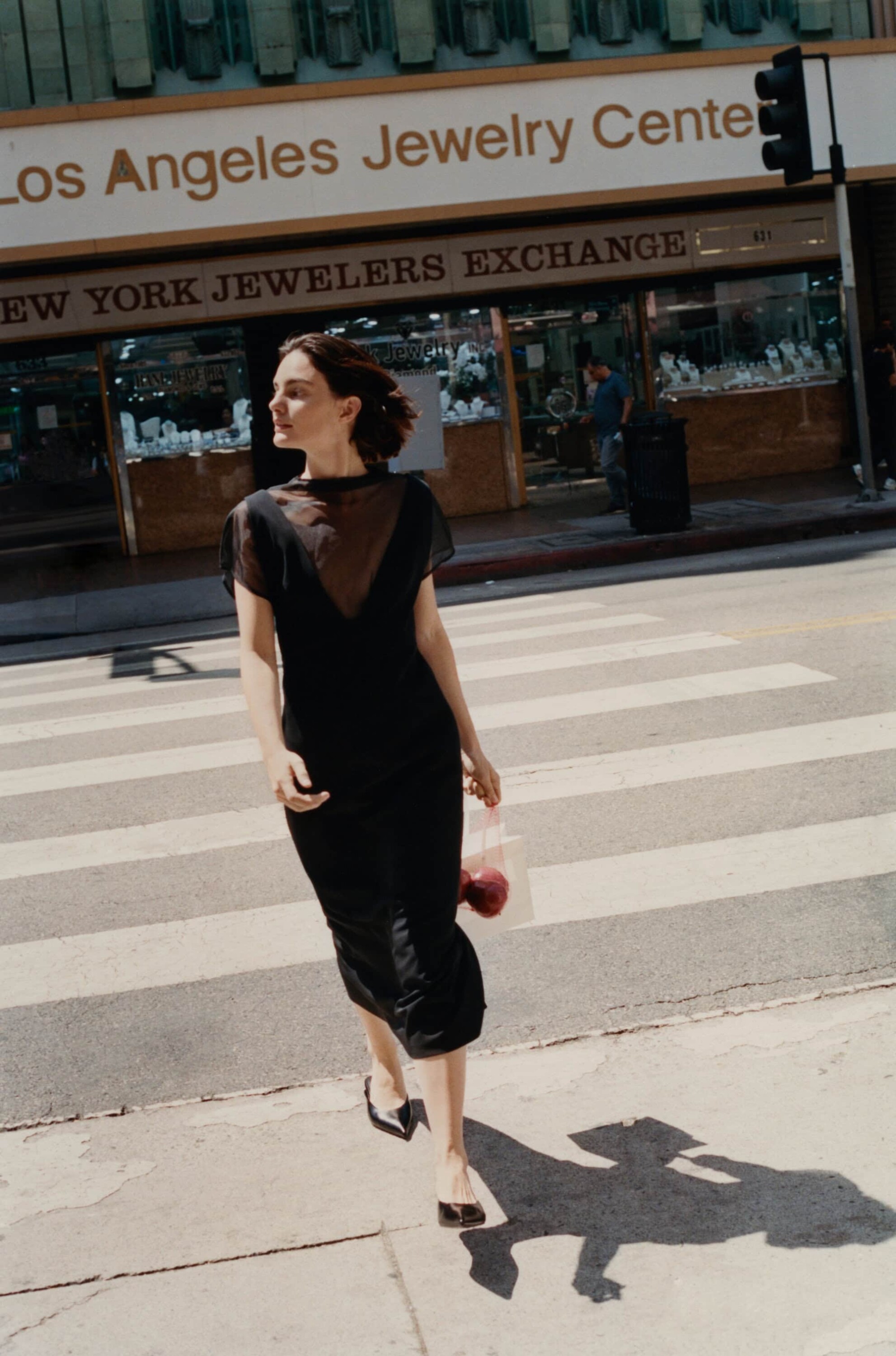 Woman in a black dress walking across a city street carrying a shopping bag past store fronts.