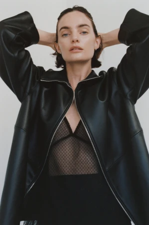Woman wearing a black leather jacket with hands behind her head against a plain light background