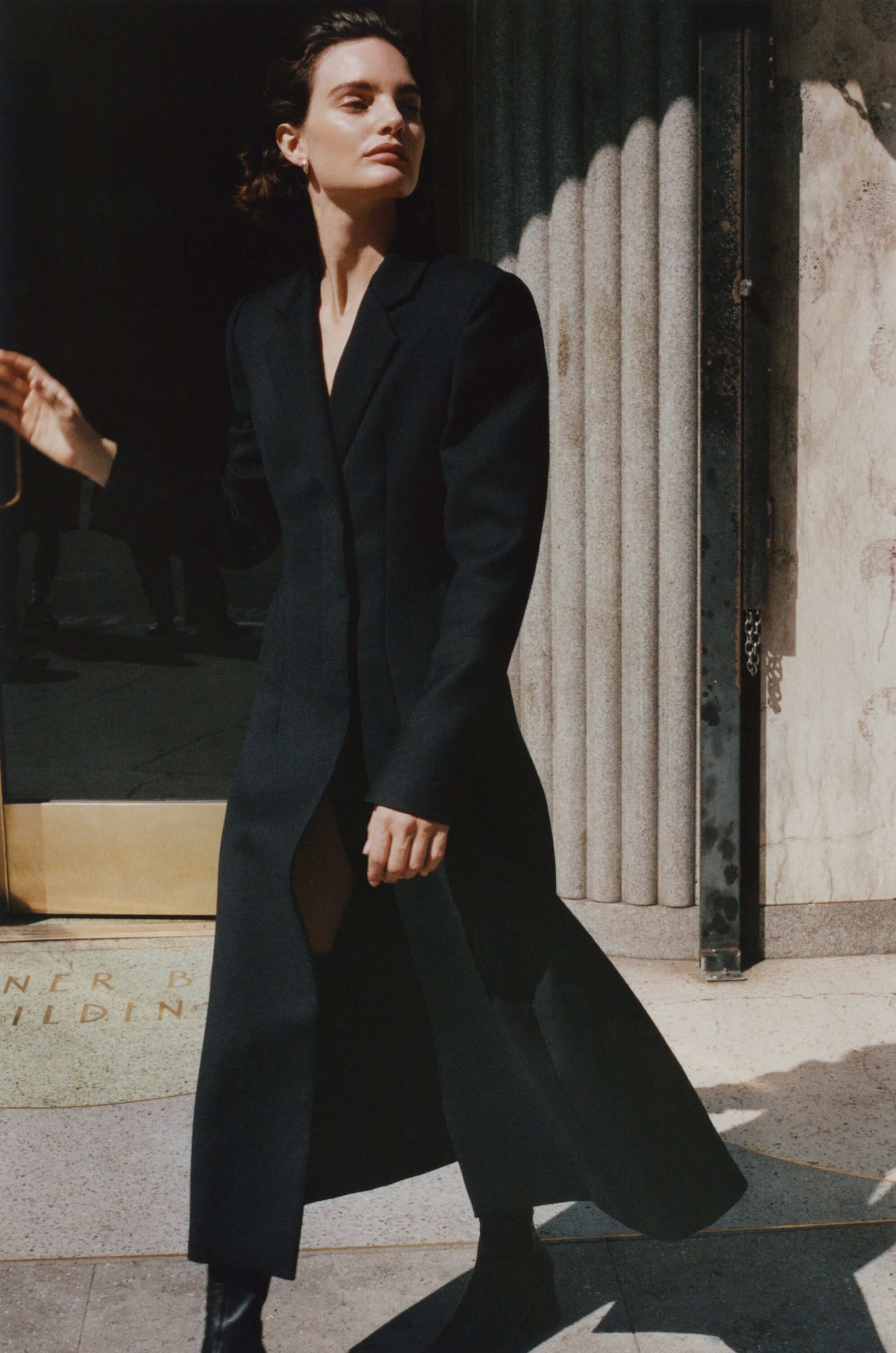 Model in a long black coat posing outside a building entrance for the Rue Sophie Fall 2024 campaign