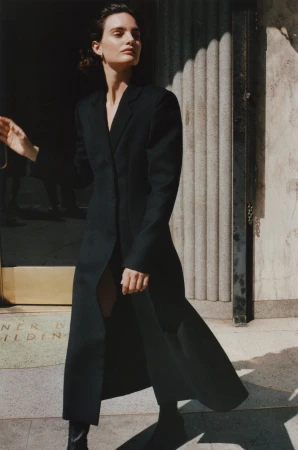 Model in a long black coat posing outside a building entrance for the Rue Sophie Fall 2024 campaign