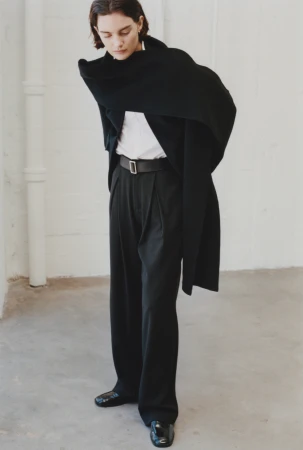 Model wearing a black coat draped over shoulders with a white shirt and black trousers against a neutral wall.
