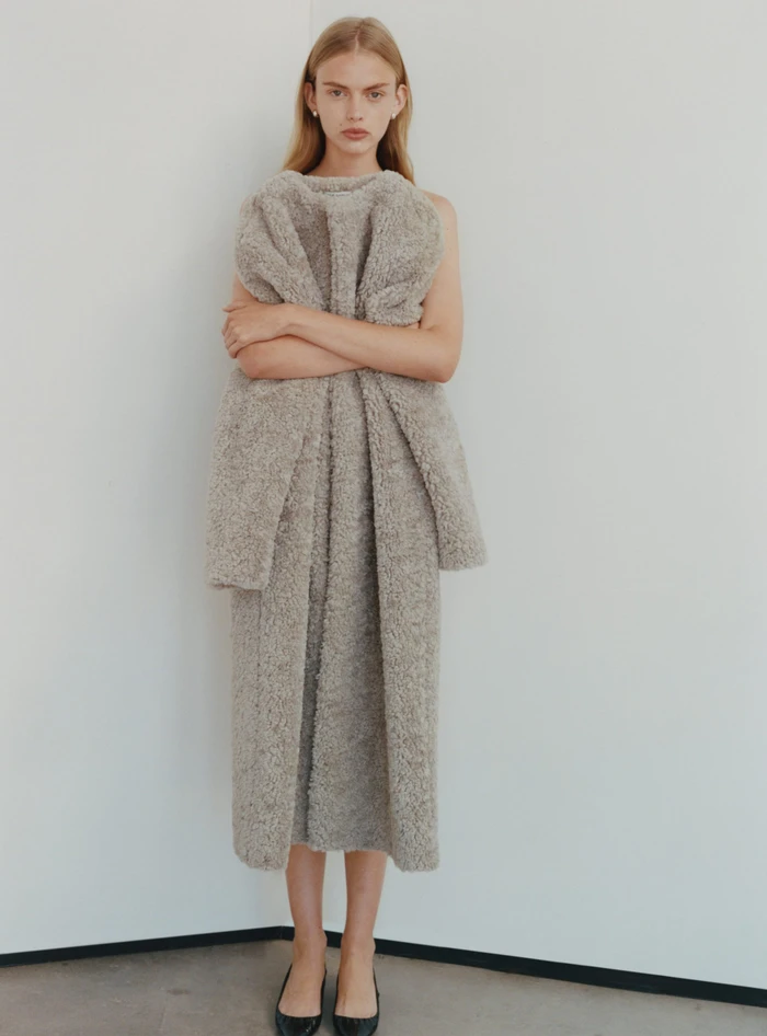 Model standing against a plain white background wearing a textured grey coat with crossed arms