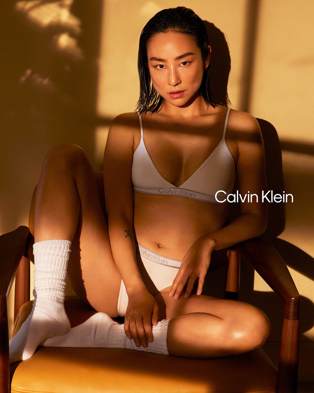Greta Lee wearing white Calvin Klein underwear and socks seated in a wooden chair with warm window light shadows
