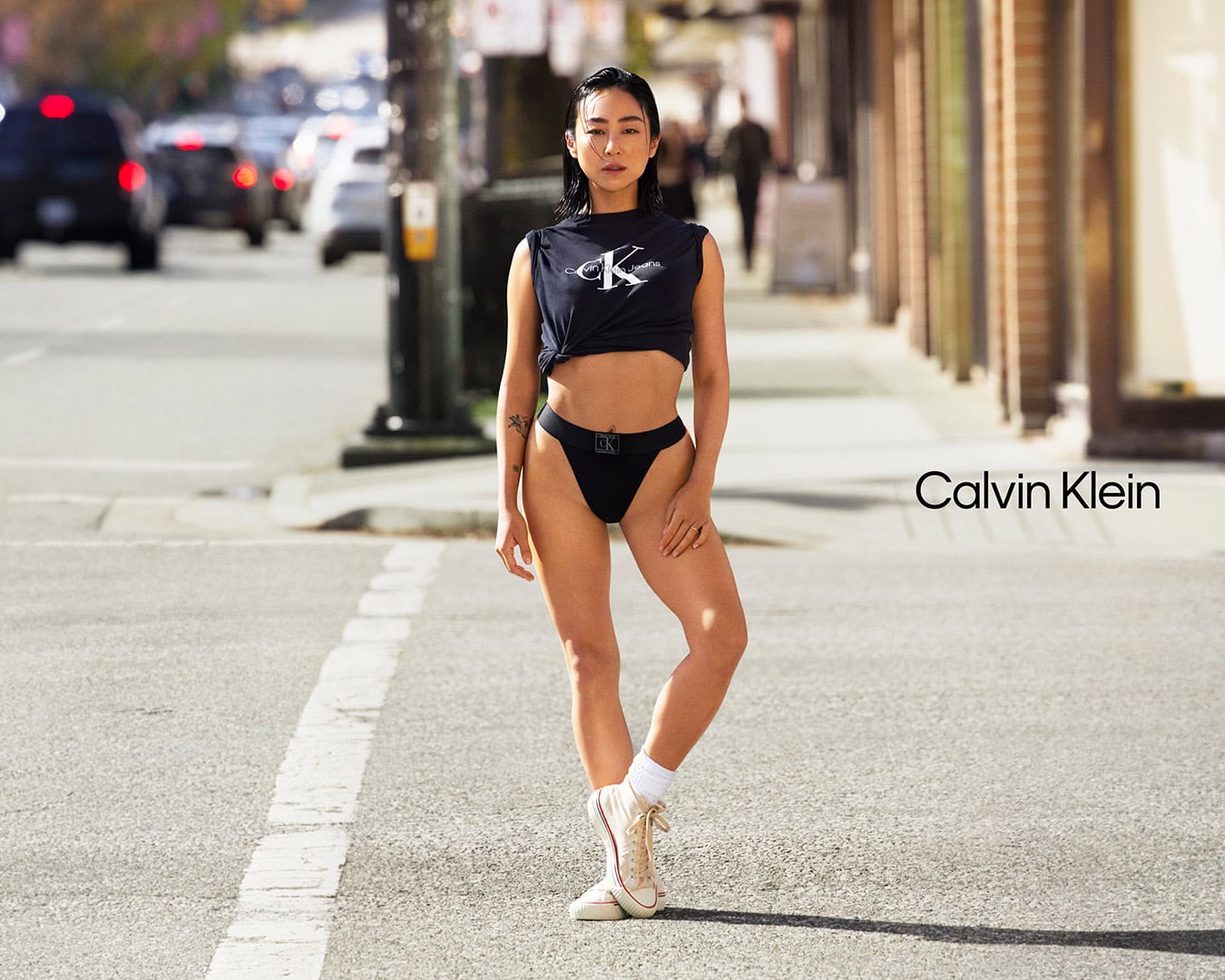 Greta Lee standing on a city street wearing a Calvin Klein black t-shirt and underwear with high top sneakers.