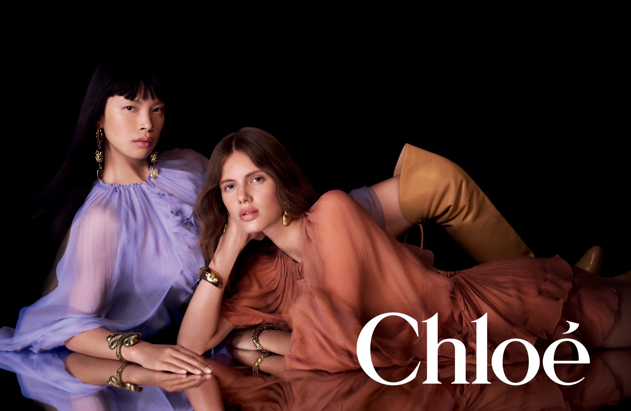 Two models wearing pastel purple and rust-coloured dresses with gold jewellery against a black background with a reflective surface.