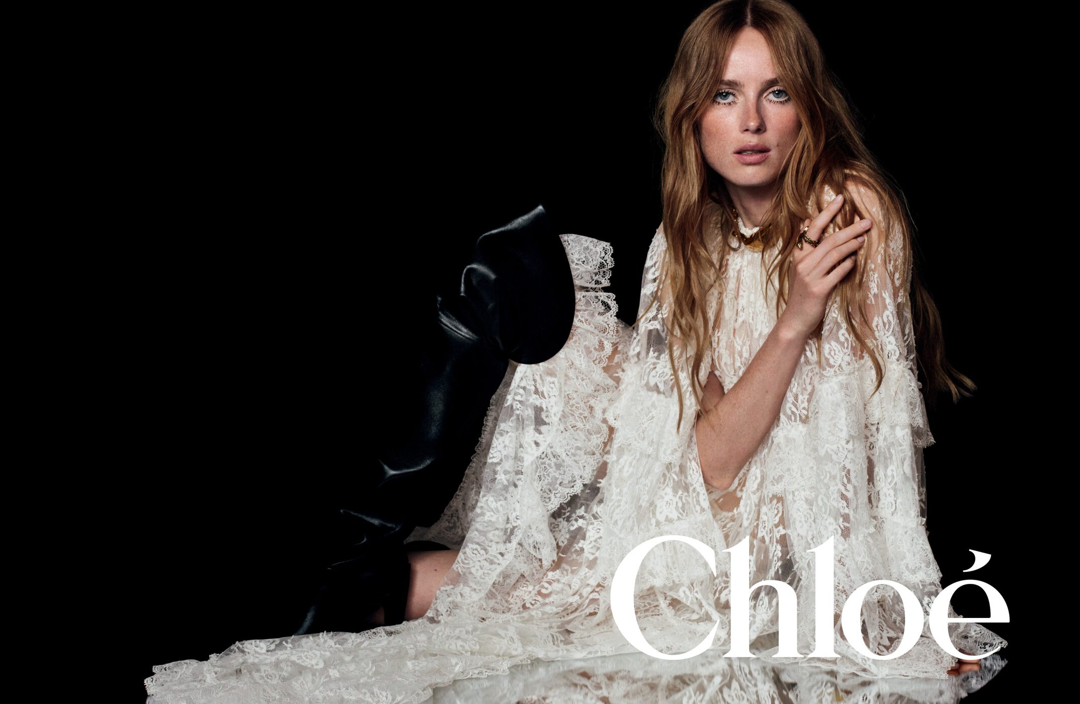 Model in white lace dress and tall black boots seated against a dark background for Chloé winter 2024 campaign