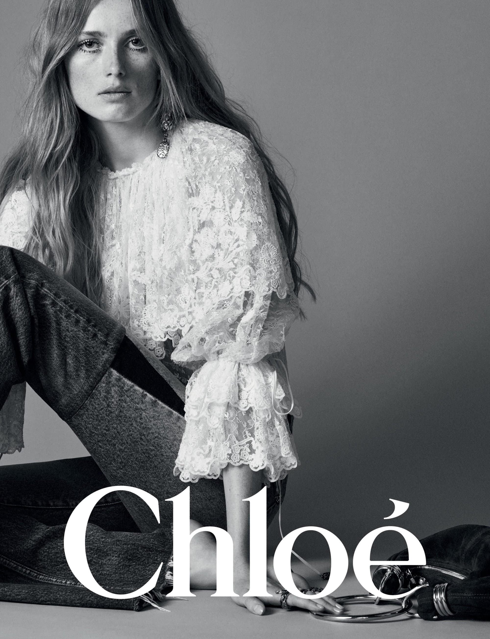 Model with long hair wearing a white lace blouse and dark jeans against a grey backdrop in the Chloé Winter 2024 campaign