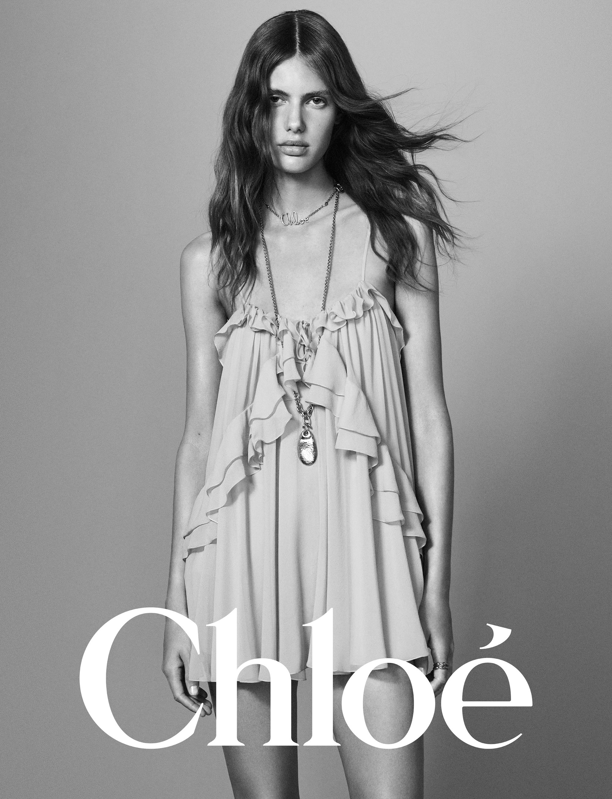 Model in a ruffled slip dress wearing layered pendant necklaces against a plain grey backdrop with bold white typography.