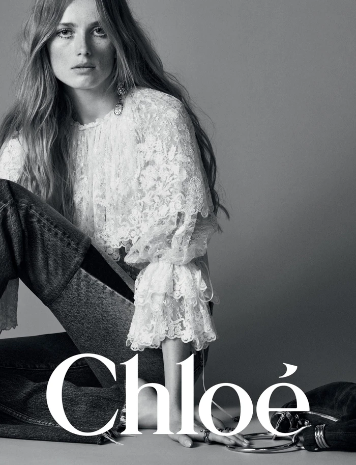 Model with long hair wearing a white lace blouse and dark jeans against a grey backdrop in the Chloé Winter 2024 campaign