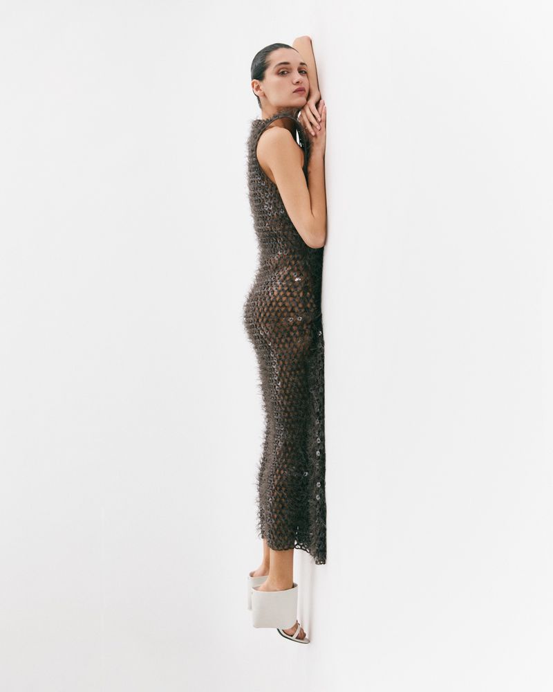 A woman leans against a white wall wearing a textured brown sleeveless dress and white block-heel sandals in a studio setting.